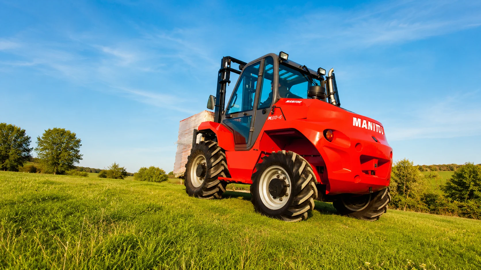 Röd Manitou-lift på grön äng under blå himmel.