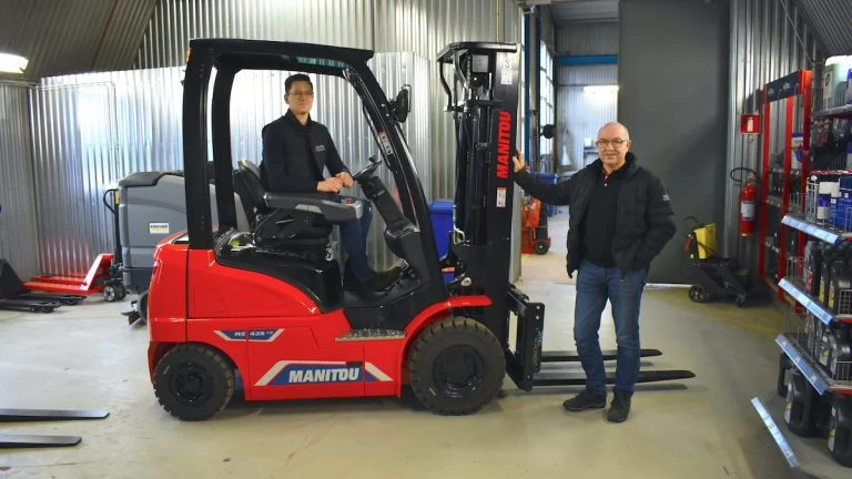 Två män står bredvid en röd Manitou-gaffeltruck inne i ett garage och visar upp Hestra Truck & Maskins expertis och löftet om en ny generation inom verkstadsutrustning. Verktyg och maskiner fyller bakgrunden.